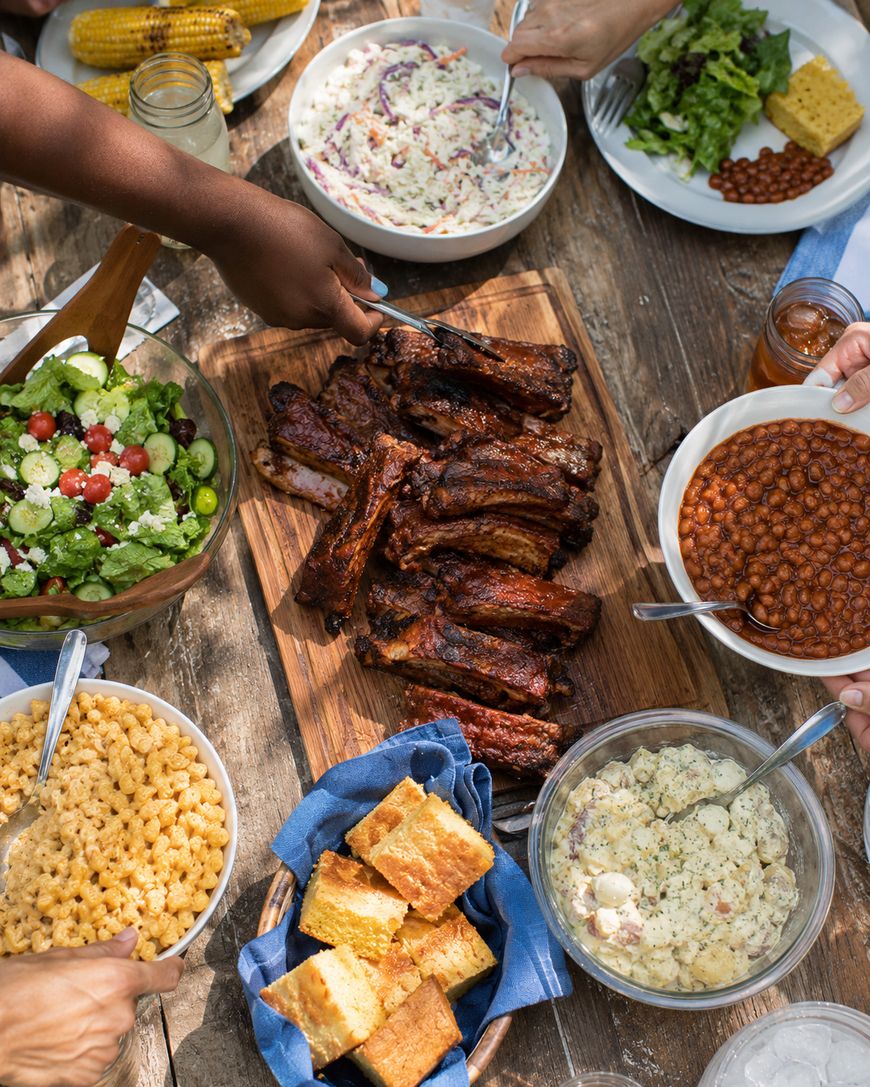 A picnic table filled with BBQ ribs, baked beans, corn on the cob, cornbread, macaroni and cheese, coleslaw, potato salad, green salad, and people serving themselves.