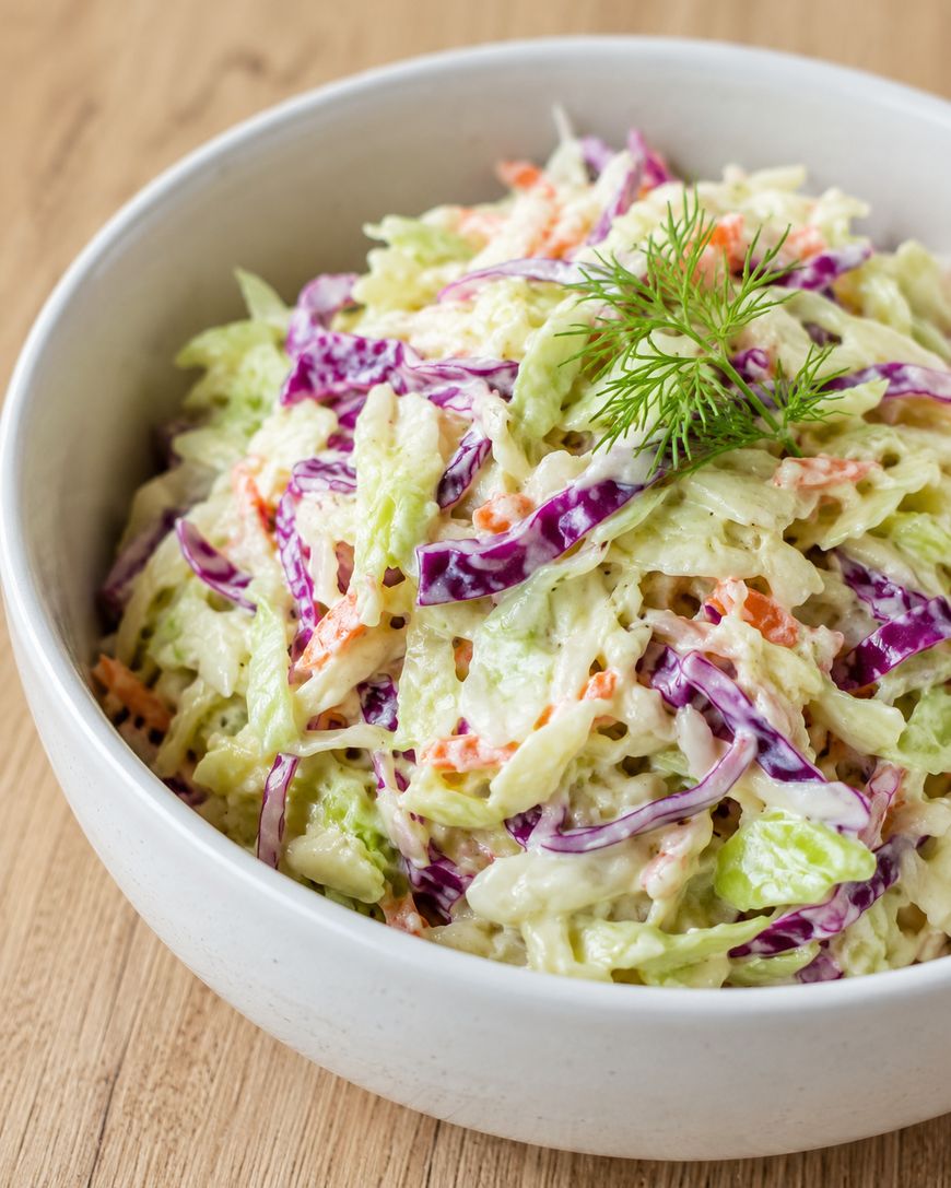 A white bowl filled with creamy coleslaw made of green and purple cabbage, carrots, and garnished with a sprig of fresh dill, placed on a wooden surface.