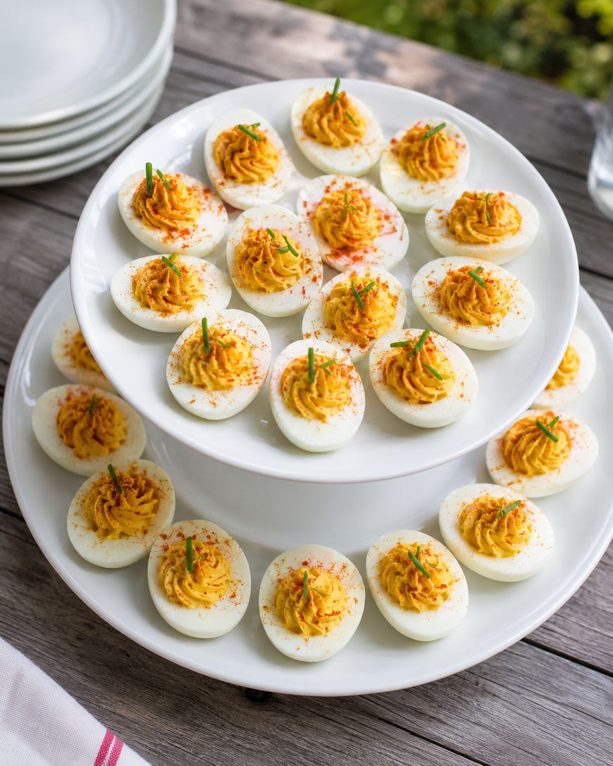 A two-tiered white serving tray displays several deviled eggs garnished with paprika and chopped chives, set on a wooden table with plates stacked in the background.