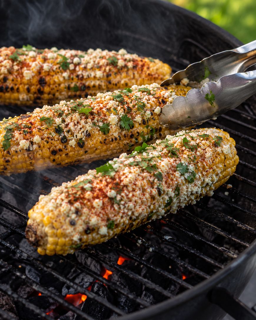 Three grilled elote (Mexican street corn) topped with cheese, chili powder, and cilantro are on a barbecue grill, with metal tongs holding one ear over glowing coals.