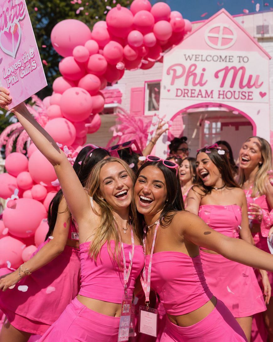 A group of smiling young women in bright pink outfits pose in front of a Welcome to Phi Mu Dream House sign, surrounded by pink balloons and decorations at a lively outdoor sorority event.