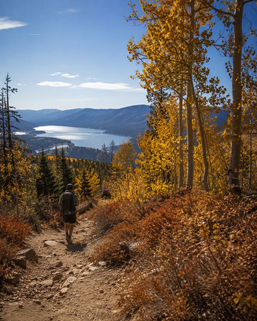 A hiker walks along a dirt trail surrounded by golden autumn trees, with a lake and mountains visible in the background under a clear blue sky.