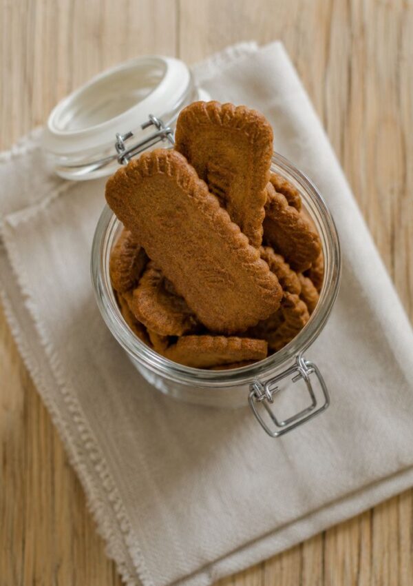 A glass jar filled with rectangular Biscoff Cookies sits on a folded beige cloth, placed on a wooden surface. The jar lid is open and rests behind the jar, showcasing this easy homemade recipe.