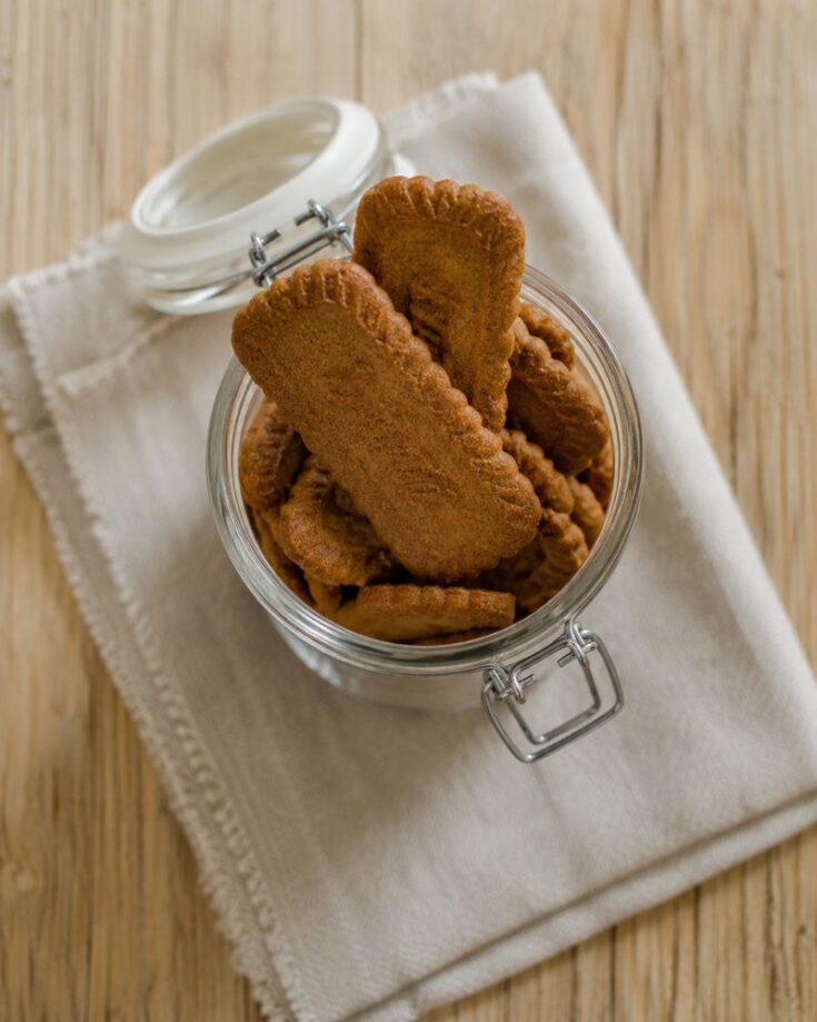 A glass jar filled with rectangular Biscoff Cookies sits on a folded beige cloth, placed on a wooden surface. The jar lid is open and rests behind the jar, showcasing this easy homemade recipe.