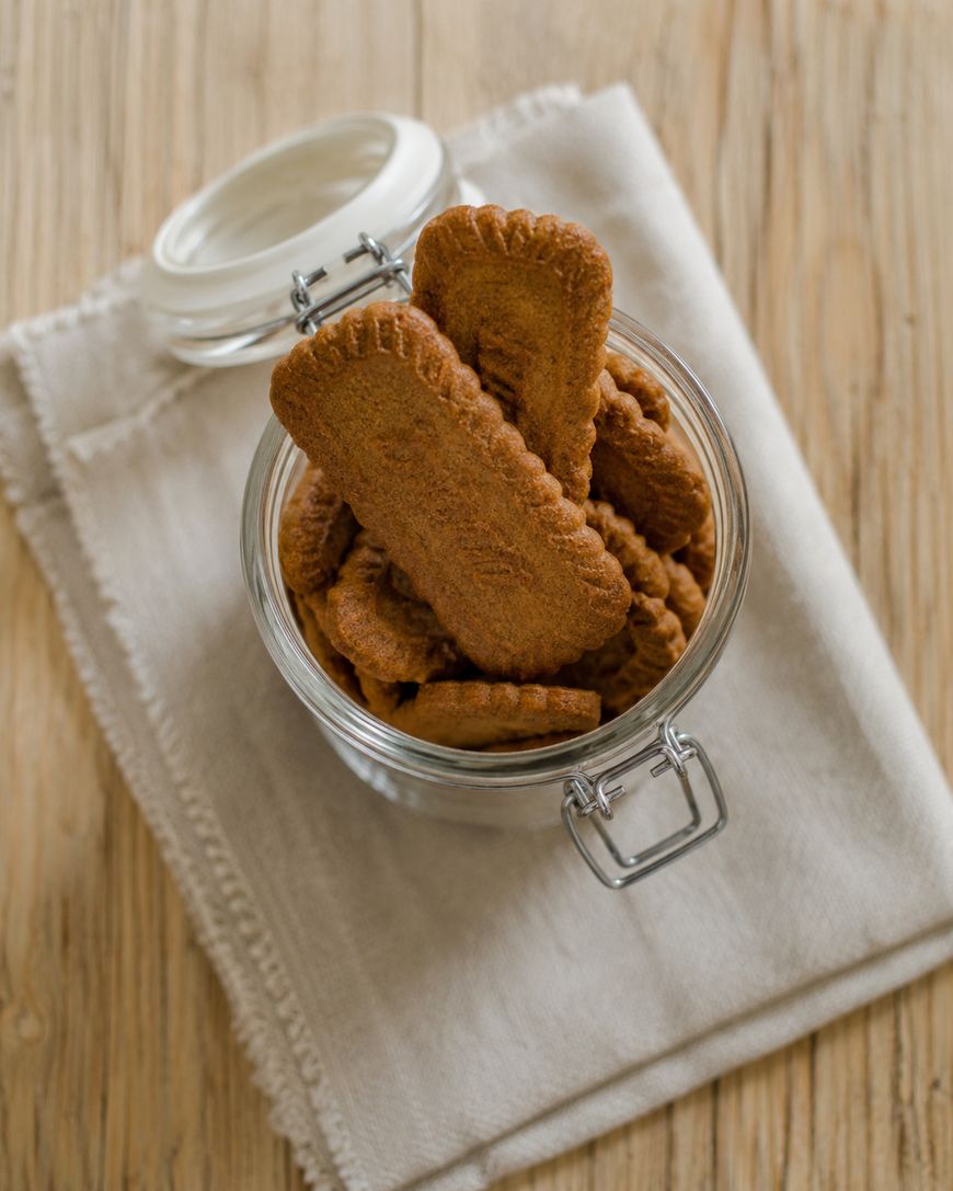 A glass jar filled with rectangular brown cookies, placed on a beige cloth napkin on a wooden surface. The jar lid is open and a few cookies are standing upright above the rim.