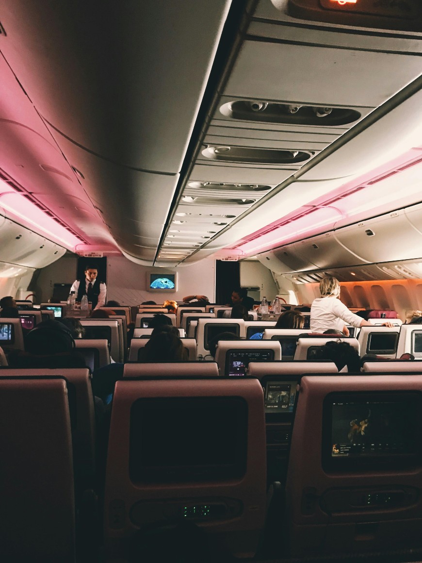 Passengers sit in an airplane cabin with pink overhead lighting. Some are watching screens, and flight attendants are moving in the aisle. The view is from the back, looking toward the front of the plane.