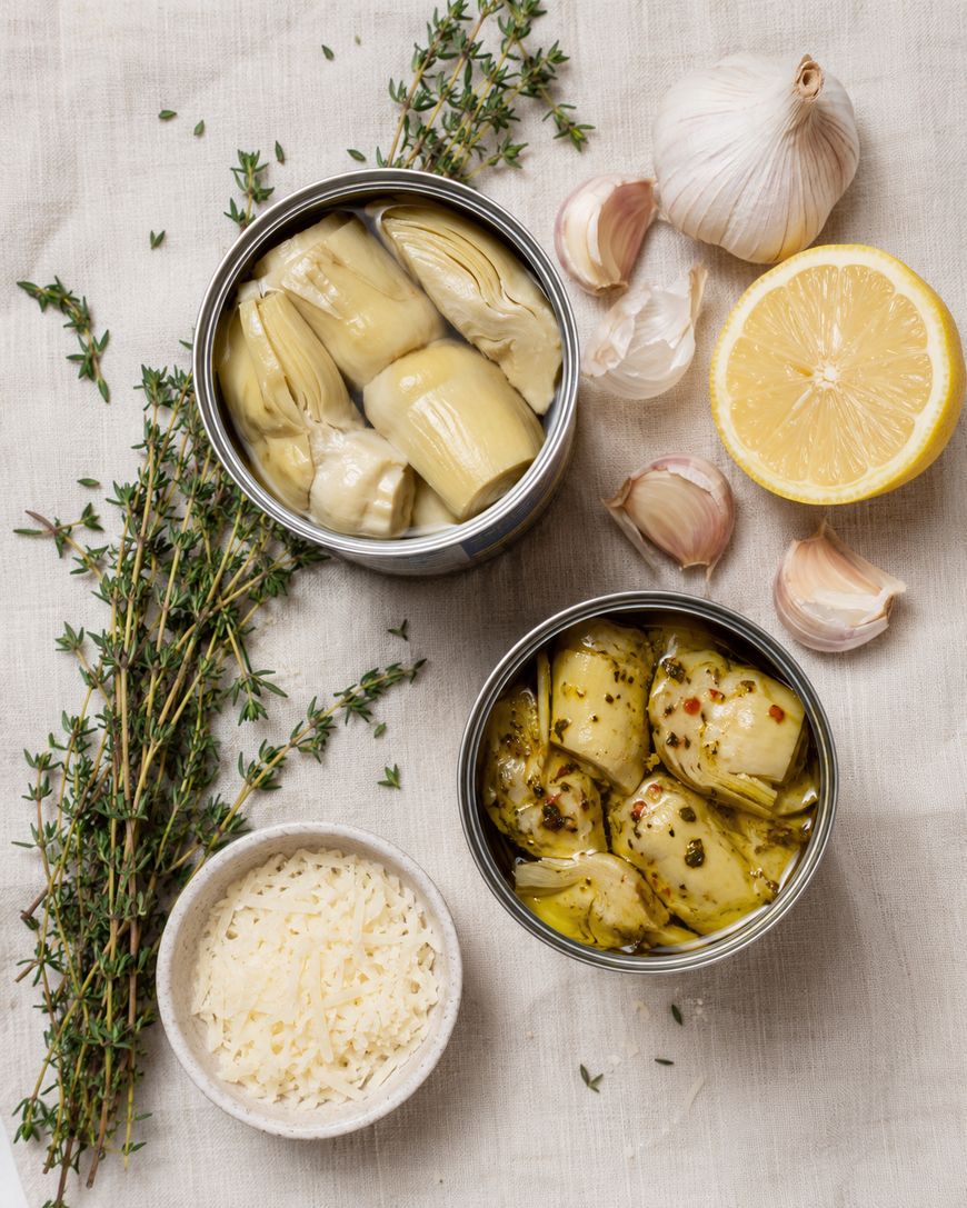 Two open cans of artichokes sit on a cloth with fresh thyme, garlic bulbs and cloves, a halved lemon, and a small bowl of grated cheese arranged around them.