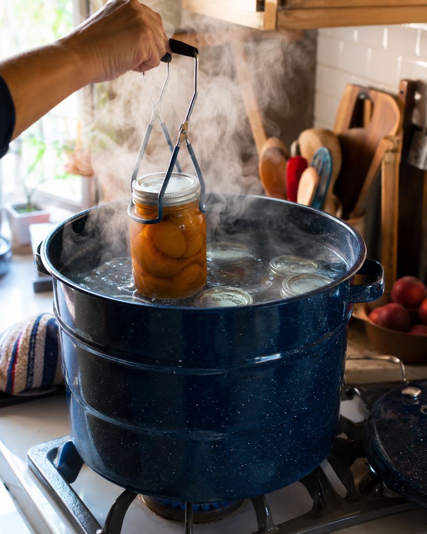 A hand uses tongs to lift a glass jar filled with peaches from a steaming blue canning pot on a stove, with other jars submerged in hot water.