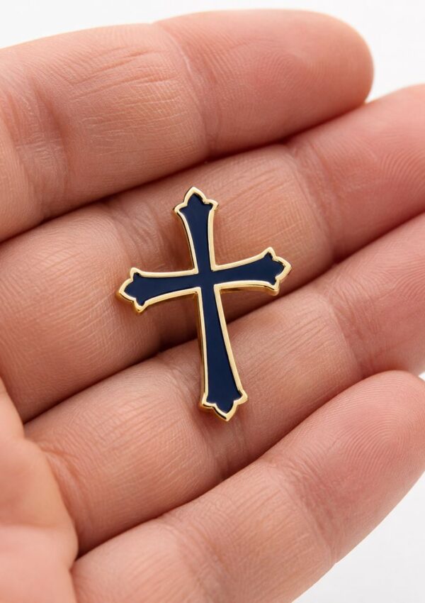 A hand holding a small pin shaped like a cross with gold edges and a dark blue interior—an elegant piece of church merchandise perfect for congregation apparel.