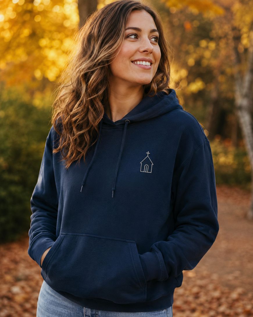 A woman with wavy brown hair, wearing a navy blue hoodie with a small church outline design, stands outdoors in a park with autumn leaves in the background, smiling and looking to the side.
