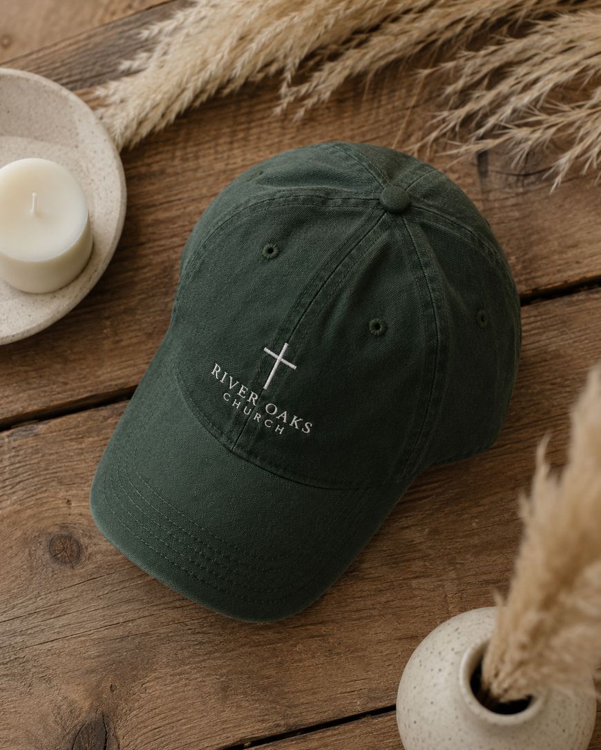 A dark green baseball cap with a white embroidered cross and the words River Oaks Church sits on a wooden surface, surrounded by a white candle, ceramic dishes, and dried pampas grass.