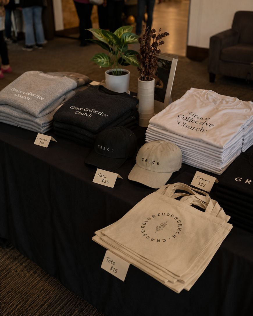 A merchandise table displays folded shirts, hats, and tote bags labeled with “Grace Collective Church” and “GRACE.” Price tags for each item are visible on a black tablecloth. A plant decorates the table.