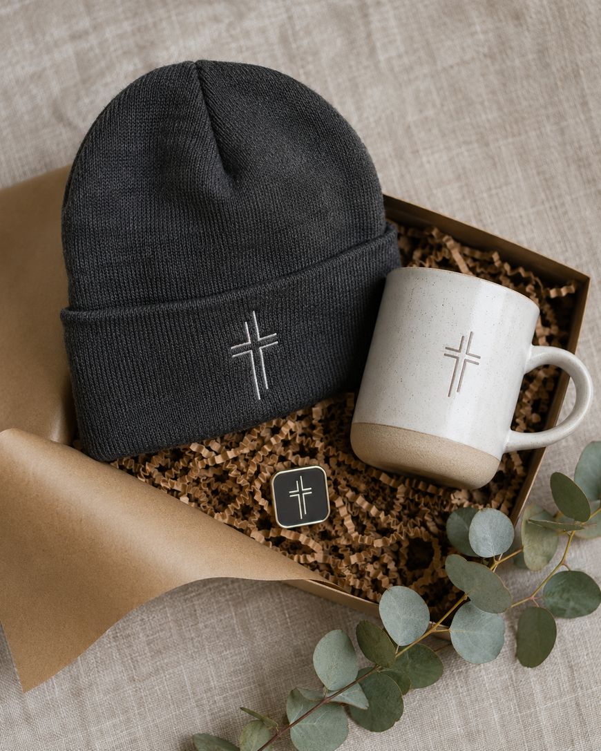 A gift box contains a gray knit beanie, a white ceramic mug, and a small pin, all featuring a simple cross design. Decorative brown paper and eucalyptus leaves surround the items.