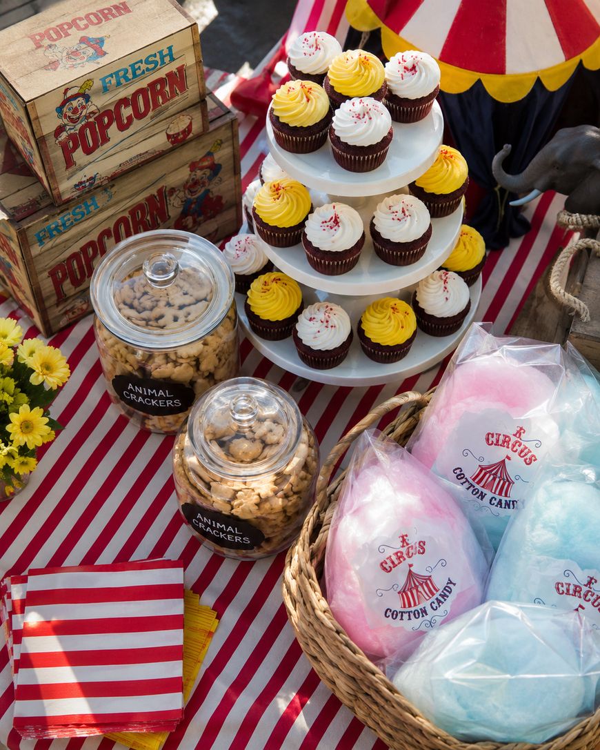 A circus-themed table with striped cloth features cupcakes, jars of animal crackers, bags of blue and pink cotton candy, popcorn boxes, and a stack of napkins, creating a festive atmosphere.