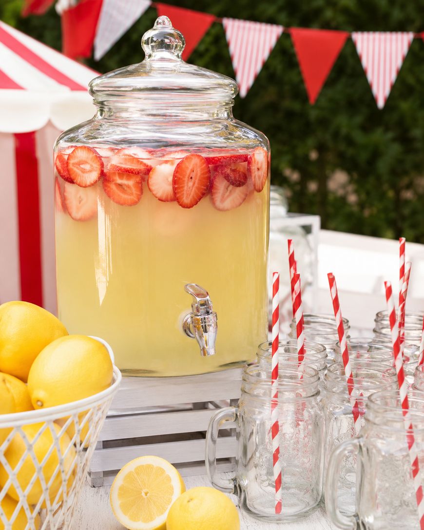 A glass drink dispenser filled with yellow lemonade and sliced strawberries sits on a table with mason jar glasses, red striped straws, fresh lemons, and festive red-and-white bunting in the background.