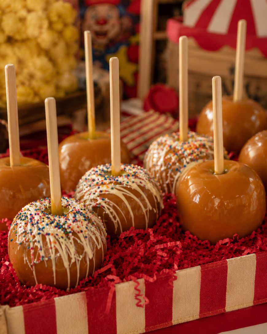 Caramel apples on sticks, some topped with white chocolate drizzle and colorful sprinkles, are displayed on red shredded paper in a festive, circus-themed setting.