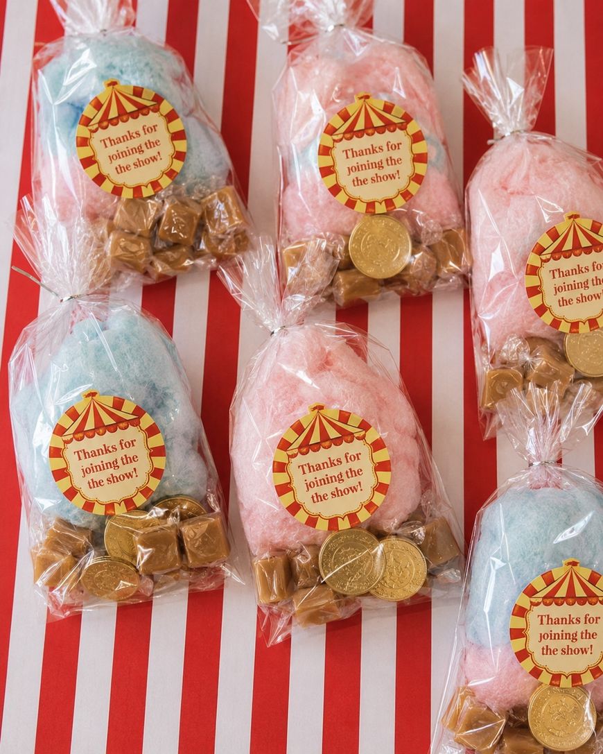 Six clear bags with pink or blue cotton candy, caramels, and gold chocolate coins, each labeled with a circus-themed sticker that says, Thanks for joining the show! on a red-and-white striped background.