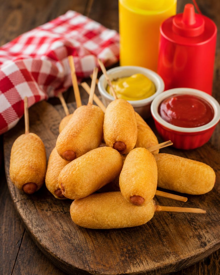A pile of mini corn dogs on wooden board with a red checkered napkin, a yellow mustard bottle, a red ketchup bottle, and small bowls of mustard and ketchup in the background.