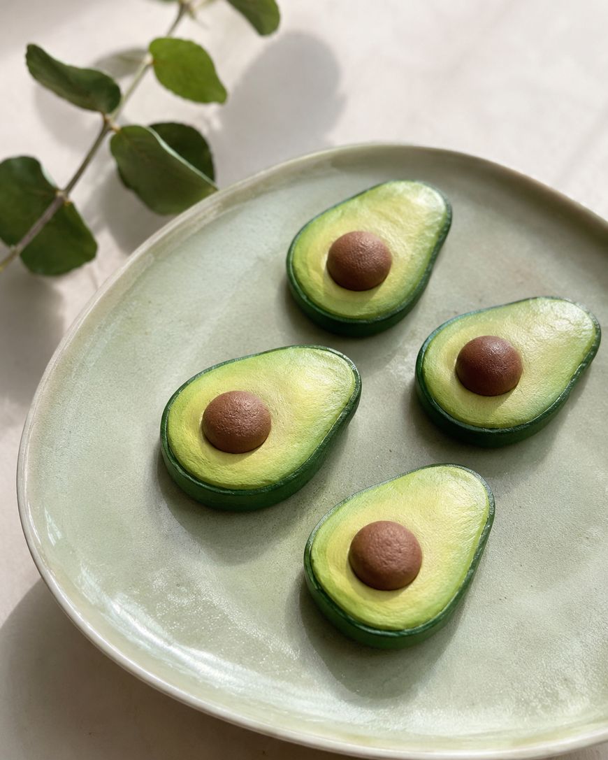 Four realistic avocado halves with brown pits are arranged on a light green ceramic plate. A eucalyptus branch lies nearby on a pale surface, adding a natural decorative touch.