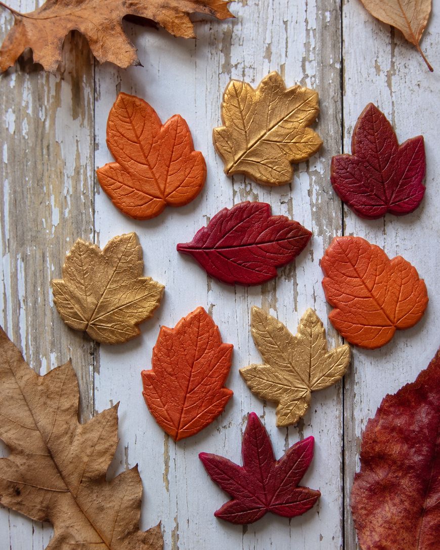 Colorful clay leaves in shades of orange, red, and gold are arranged on a rustic, weathered wooden surface, surrounded by real dried brown leaves.