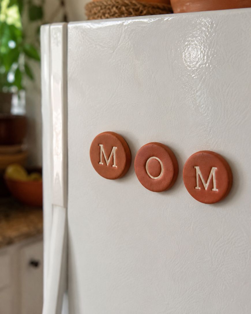 Three round, brown magnets with white letters spelling MOM are arranged on a white refrigerator in a cozy kitchen setting.
