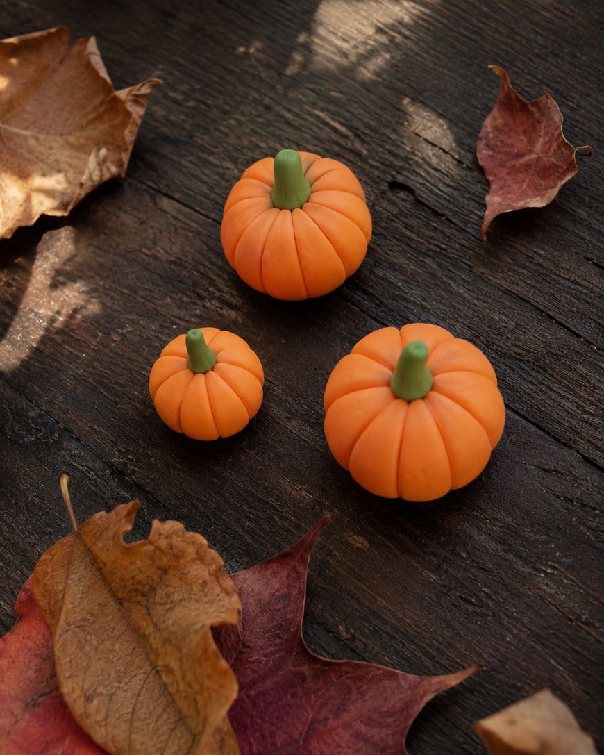 Three small orange pumpkins with green stems are arranged on a dark wooden surface, surrounded by scattered dry autumn leaves in shades of brown and red.
