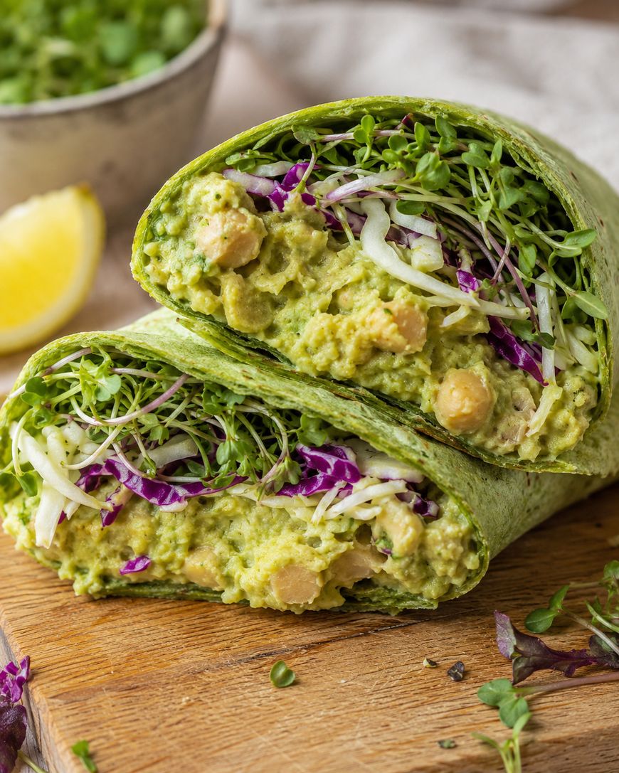 A spinach wrap filled with chickpeas, creamy avocado spread, shredded red and green cabbage, and fresh sprouts, cut in half and displayed on a wooden board with a lemon wedge in the background.