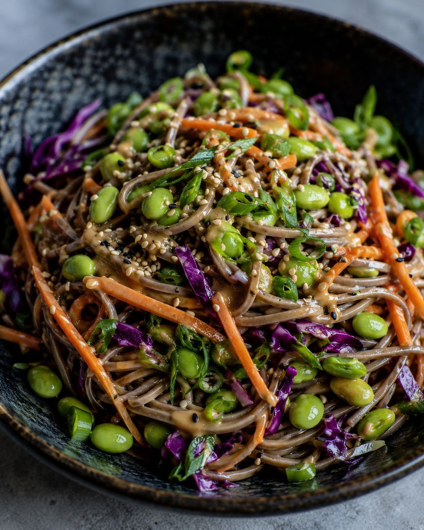 A bowl of soba noodles mixed with shredded purple cabbage, julienned carrots, edamame, chopped green onions, and sesame seeds, all topped with a creamy dressing.