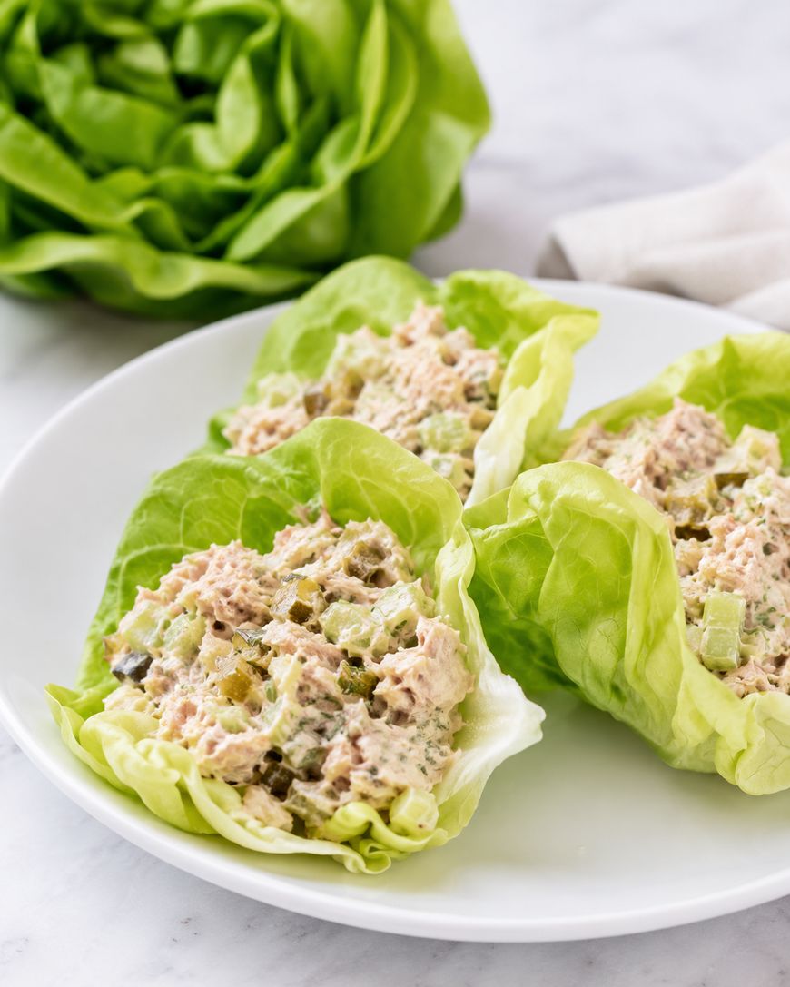 Three lettuce leaves filled with tuna salad, containing chopped celery and pickles, are arranged on a white plate. A head of lettuce and a napkin are in the background on a marble surface.