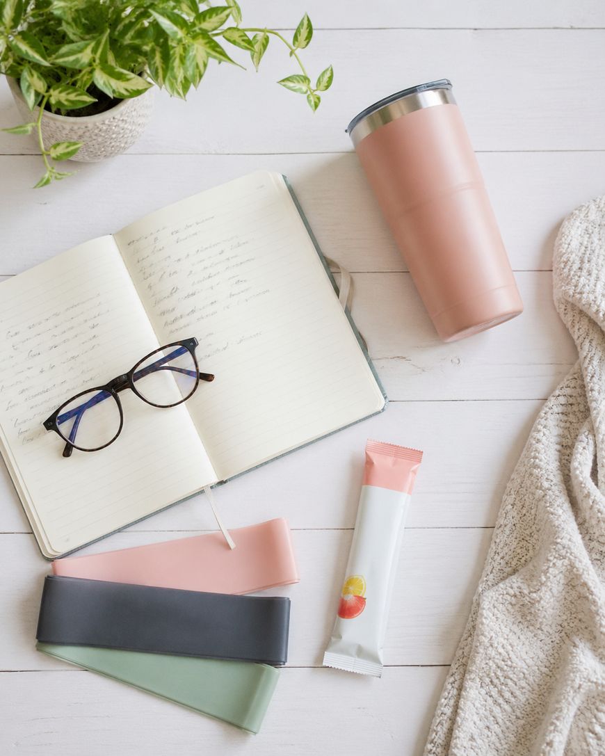 Flat lay of an open notebook with handwritten notes, a pair of glasses, resistance bands, a pink tumbler, an energy bar, a beige knit blanket, and a potted plant on a white wooden surface.