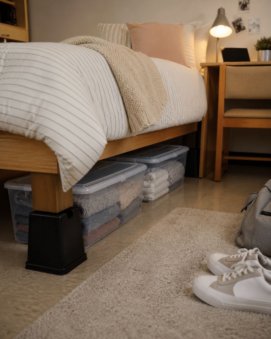 A neatly made bed with striped bedding is raised on bed risers, with clear storage bins filled with folded clothes underneath. A desk, chair, lamp, and sneakers on a rug are also visible in the cozy room.