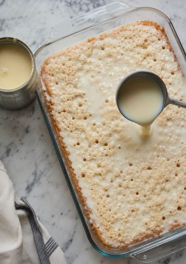 A glass baking dish with a sheet cake is being soaked with sweetened condensed milk from a ladle—perfect for condensed milk recipes and sweet treats. A can of milk and a kitchen towel rest nearby on a marble surface.