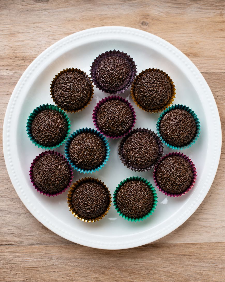 A white plate holds thirteen chocolate brigadeiros in colorful paper cups, arranged in a circular pattern on a wooden surface.