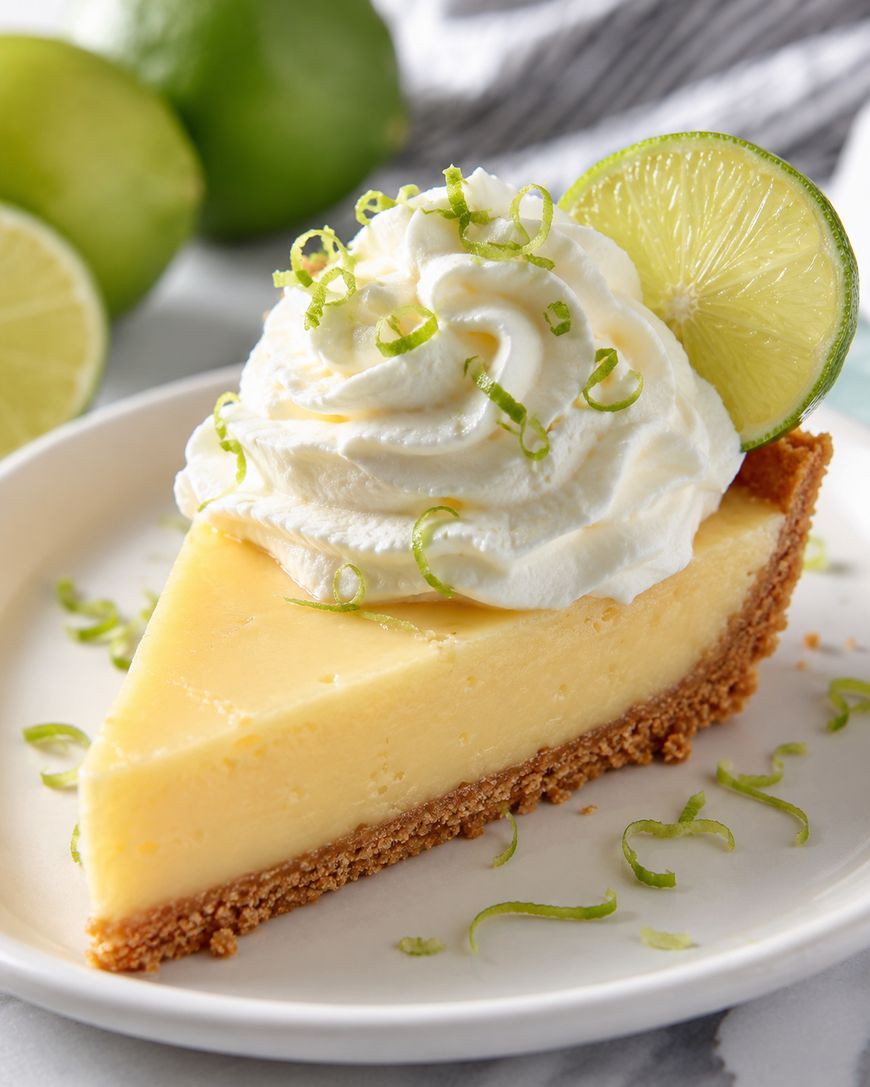 A slice of key lime pie with a graham cracker crust, topped with whipped cream, lime zest, and a slice of lime, served on a white plate with limes in the background.