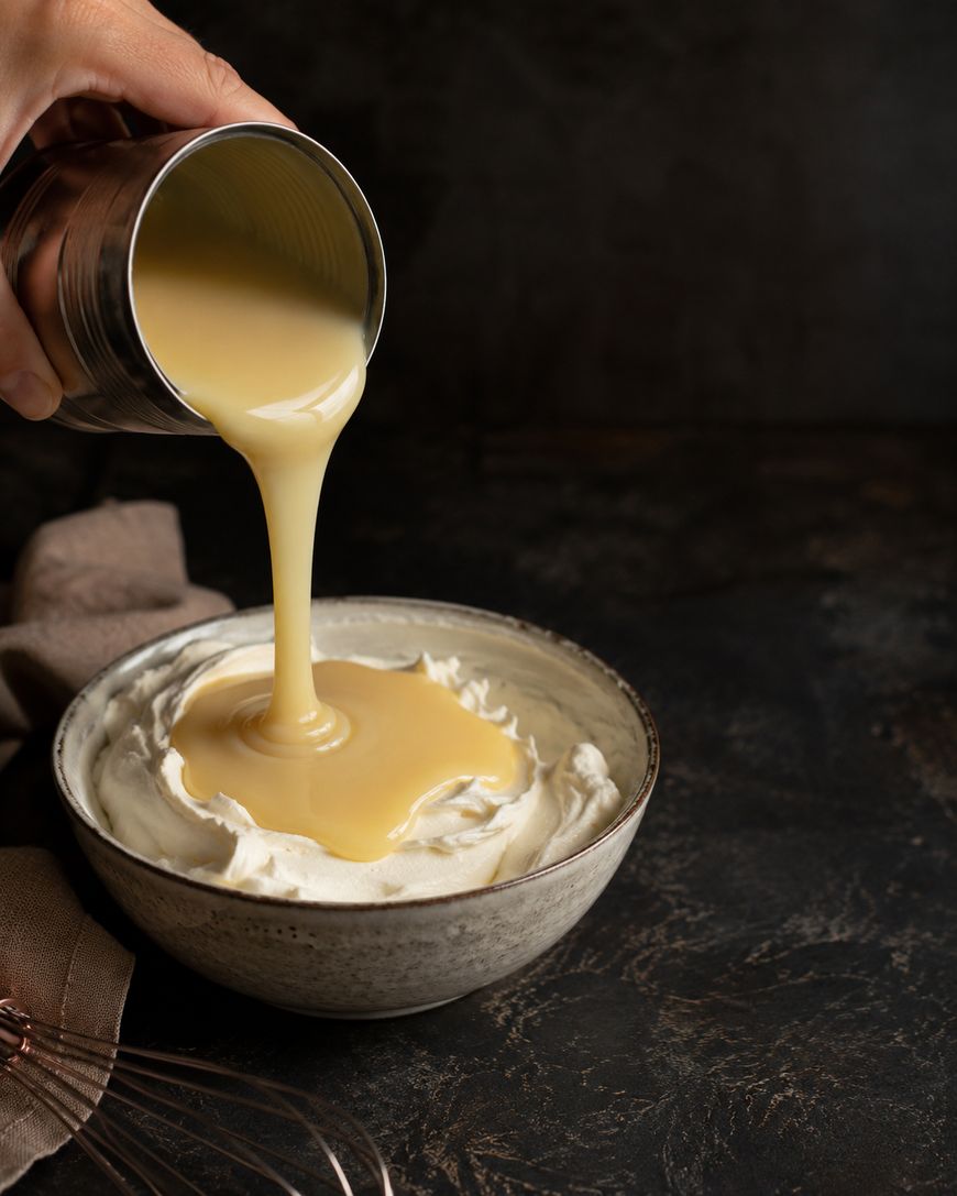 A hand pours sweetened condensed milk from a can onto a bowl of whipped cream, with a whisk and a brown cloth nearby on a dark surface.