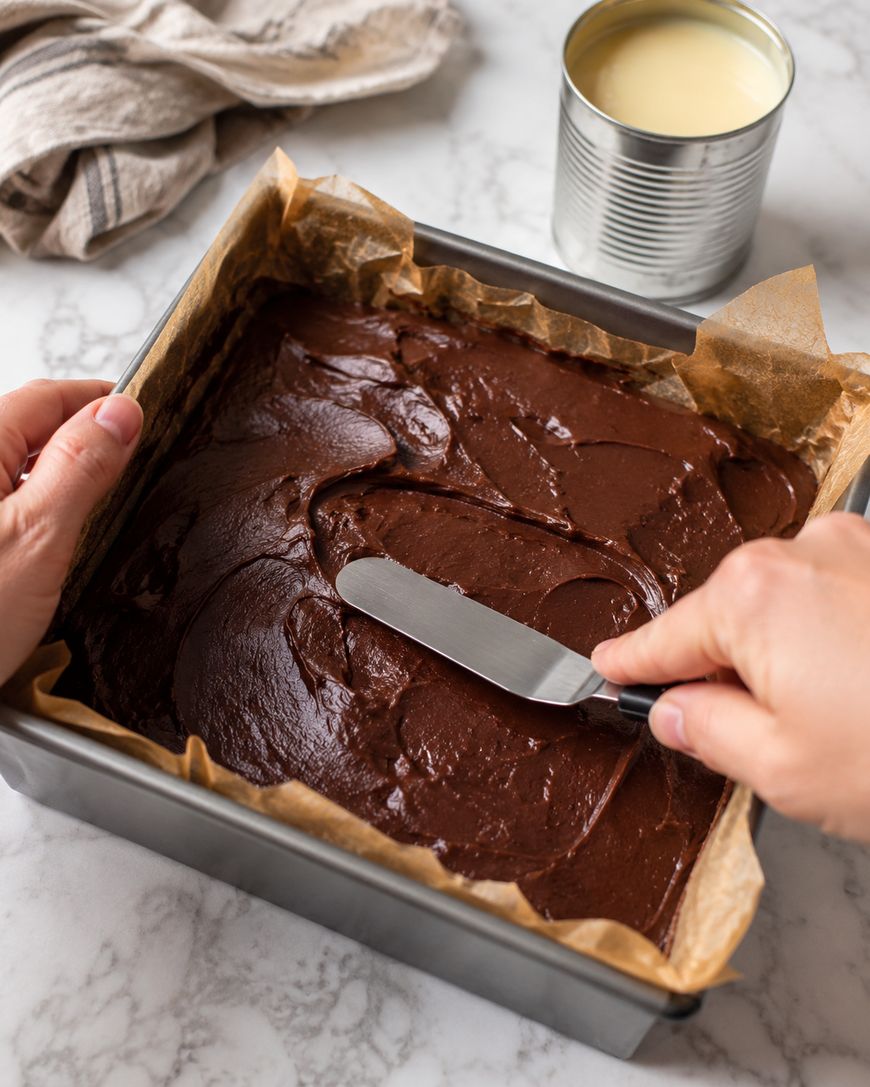 A person spreads chocolate batter in a parchment-lined square baking pan with an offset spatula. An open can of condensed milk and a kitchen towel are on a marble countertop nearby.