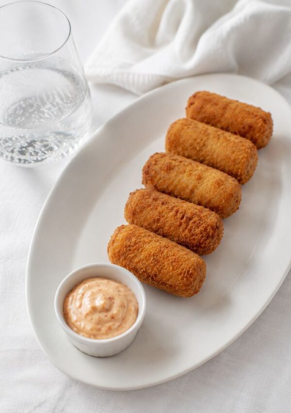 Five golden-brown croquettes, inspired by classic Cuban recipes, are arranged in a row on a white oval plate with a small bowl of dipping sauce and a glass of water nearby, set on a white tablecloth.