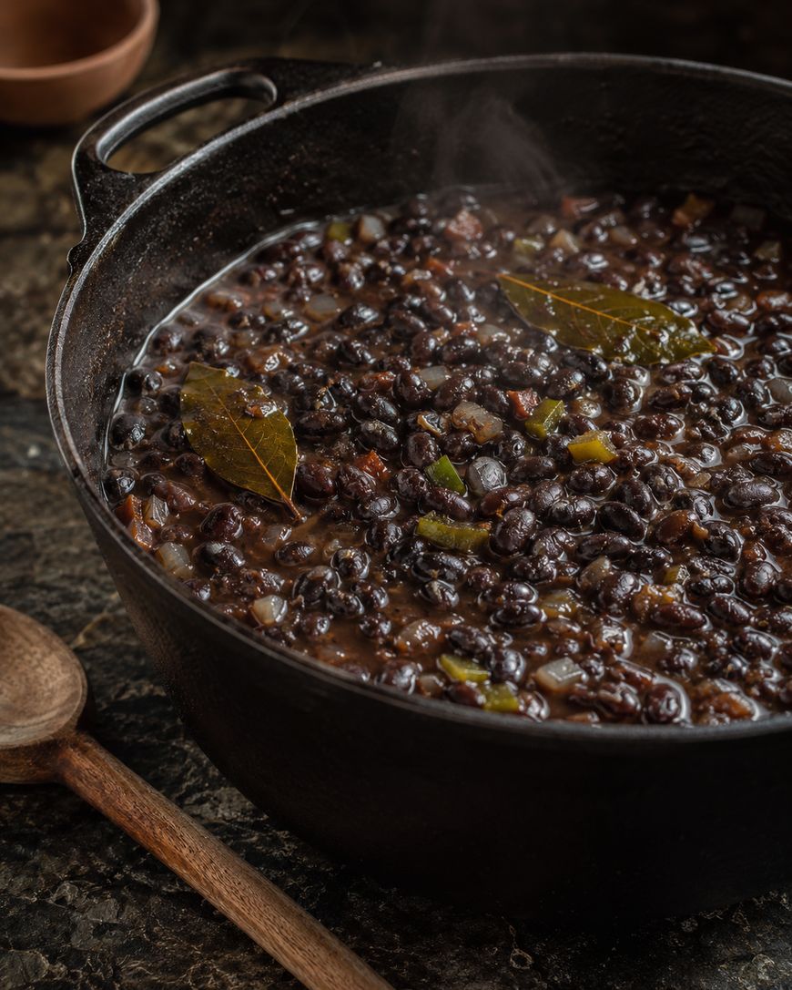 A steaming cast iron pot filled with cooked black beans, diced vegetables, and bay leaves sits on a dark surface next to a wooden spoon.