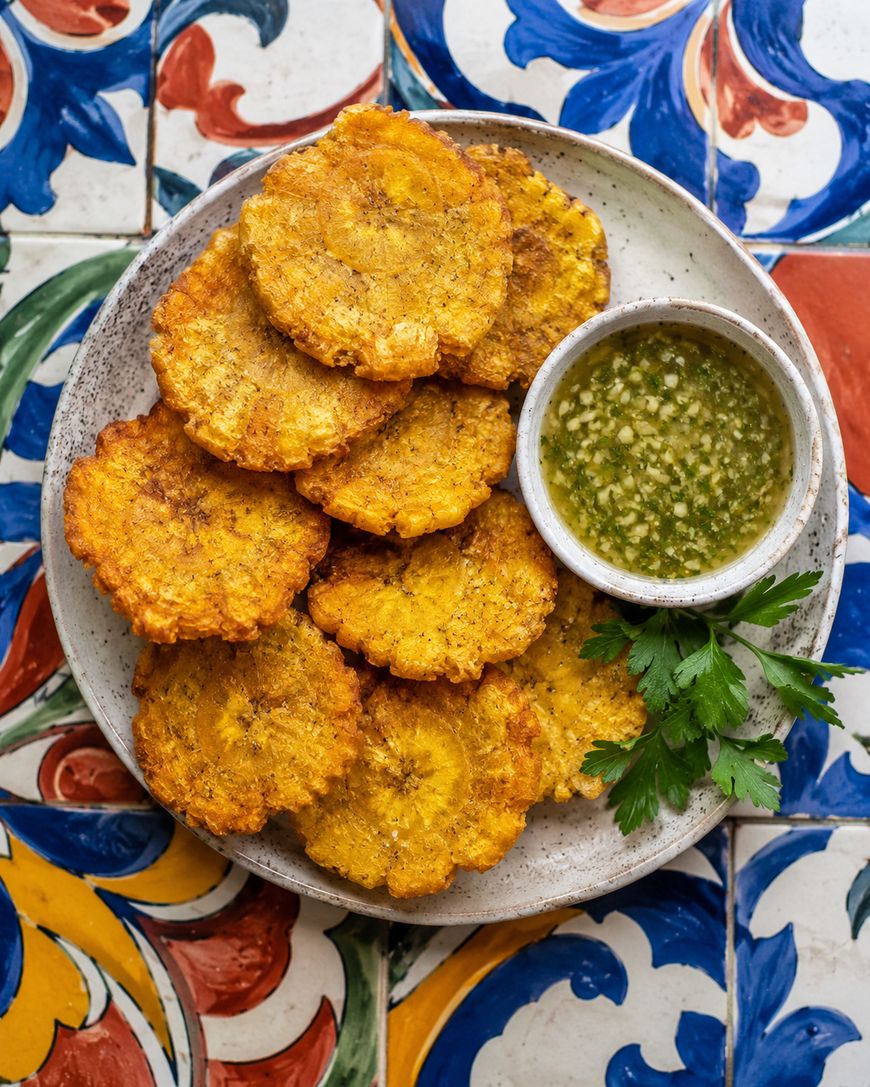 A plate of golden, crispy tostones (fried plantains) arranged around a small bowl of green dipping sauce, garnished with parsley, sits on a colorful, patterned tile surface.