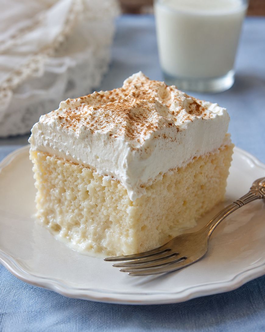 A slice of moist tres leches cake topped with whipped cream and a sprinkle of cinnamon sits on a white plate with a fork; a glass of milk is in the background.
