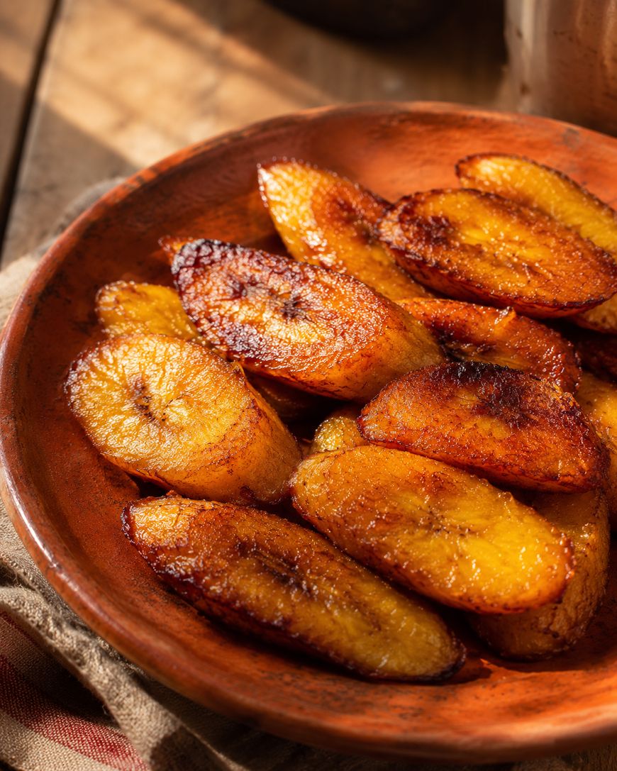 A clay plate filled with golden-brown, fried plantain slices sits on a rustic wooden surface, with warm sunlight highlighting their caramelized edges.