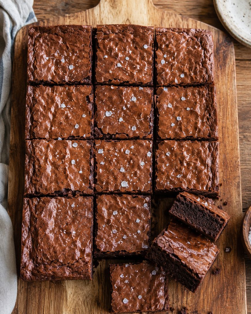 A tray of fudgy brownies cut into squares on a wooden board, with a few pieces stacked and a sprinkling of sea salt on top.