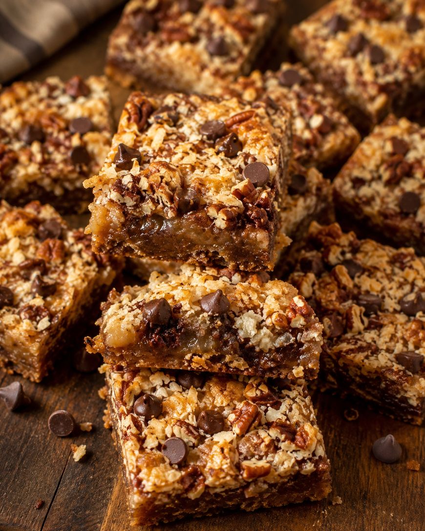 A close-up of several gooey pecan pie bars topped with chocolate chips and chopped pecans, stacked on a wooden surface. The bars have a crumbly, golden-brown crust and rich, caramelized filling.