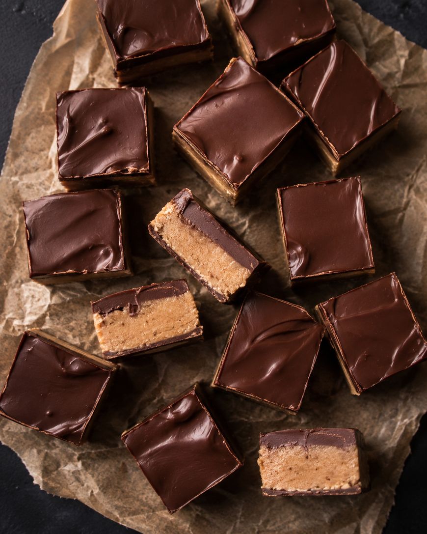 Squares of chocolate-covered peanut butter bars are arranged on crumpled brown parchment paper. Some bars are cut to reveal a thick peanut butter filling beneath a smooth chocolate layer.