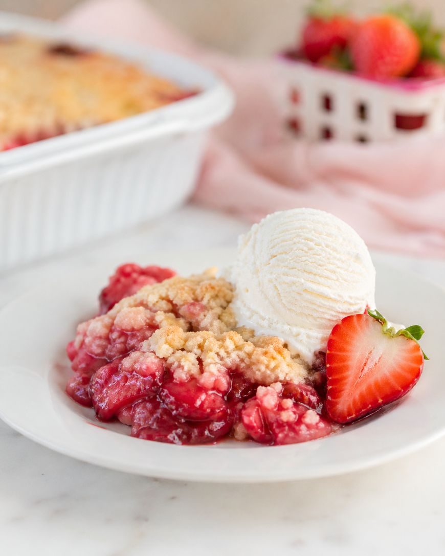 A plate of strawberry crumble topped with a scoop of vanilla ice cream and a halved strawberry. In the background, a basket of fresh strawberries and a baking dish are visible.