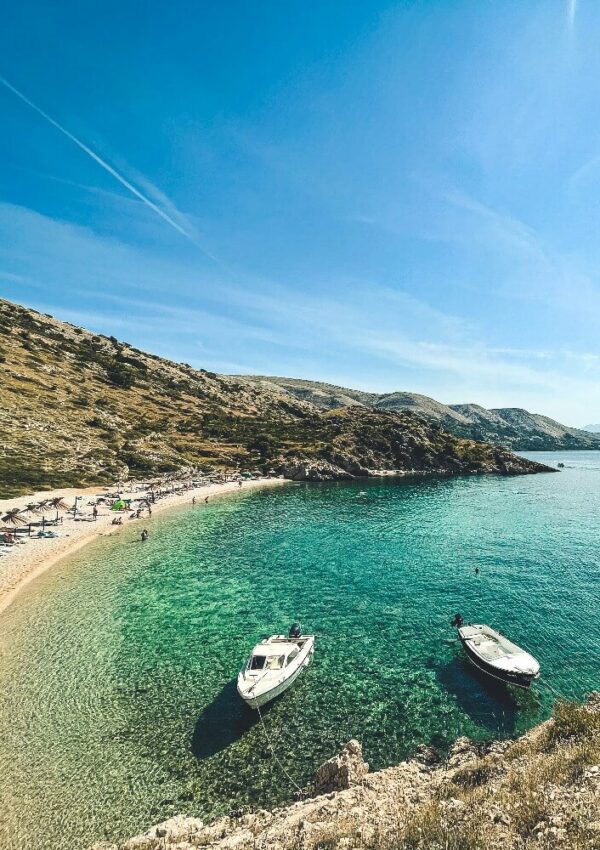 A small, curved beach with clear turquoise water and two boats anchored near the shore, surrounded by rocky hills under a bright blue sky. People relax under umbrellas along the sandy beach.