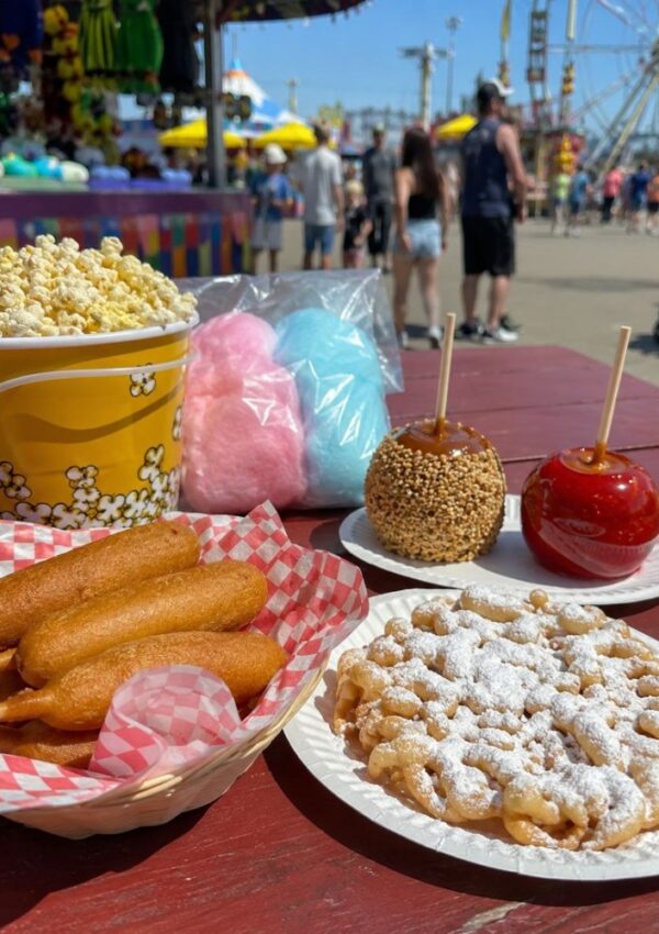 A table at a fair displays funnel cake, caramel and candy apples, cotton candy, popcorn, and corn dogs—classic fair food recipes you can make at home to taste like the real thing. In the background, people walk by colorful booths with a Ferris wheel and rides visible.