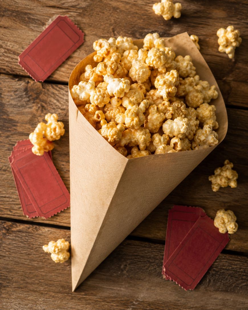 A paper cone full of caramel popcorn sits on a wooden surface, surrounded by scattered pieces of popcorn and several red admit-one tickets.