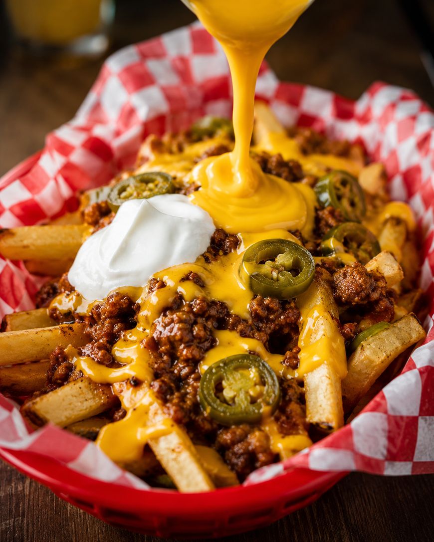 Basket of loaded fries topped with ground beef, sliced jalapeños, melted cheese sauce, and a dollop of sour cream, with more cheese sauce being poured over the top.