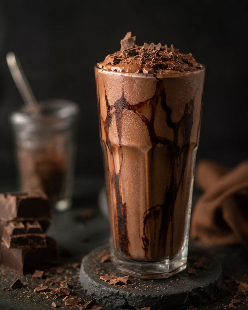 A tall glass of chocolate milkshake topped with chocolate shavings and drizzled with chocolate syrup, set on a dark surface with chunks of chocolate and a blurred jar in the background.